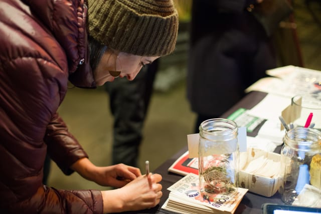 Woman in a brown puffer jacket and knit hat writing at a table during the Love Bomb 2017 event, surrounded by promotional materials and donation jars supporting My Voice Music.